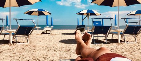 Entspannt übereinanderliegende Beine in Badeshorts an einem Strand mit vielen Strandliegen un Sonnenschirmen